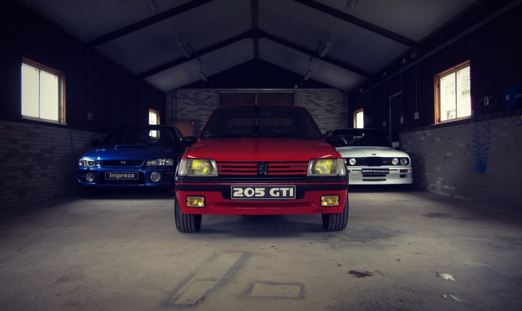 Peugeot 205 GTI frontaal in een donkere schuur, met een Subaru Impreza en een BMW M3 E30 ernaast