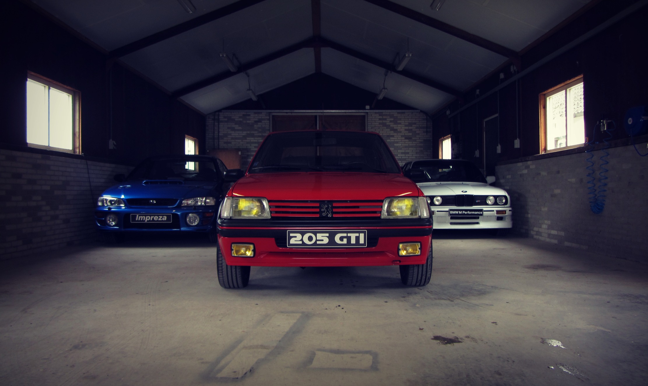 Peugeot 205 GTI frontaal in een donkere schuur, met een Subaru Impreza en een BMW M3 E30 ernaast