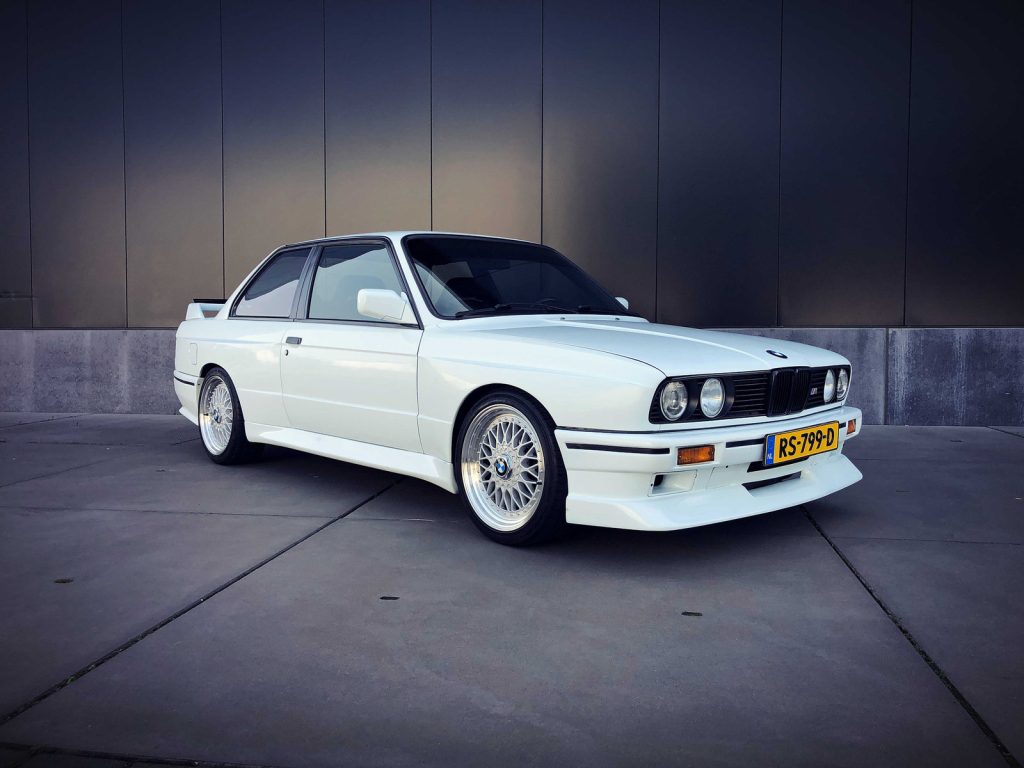 BMW M3 Tribute 2.5 (E30), driekwart van voren tegen een donkere gevel
